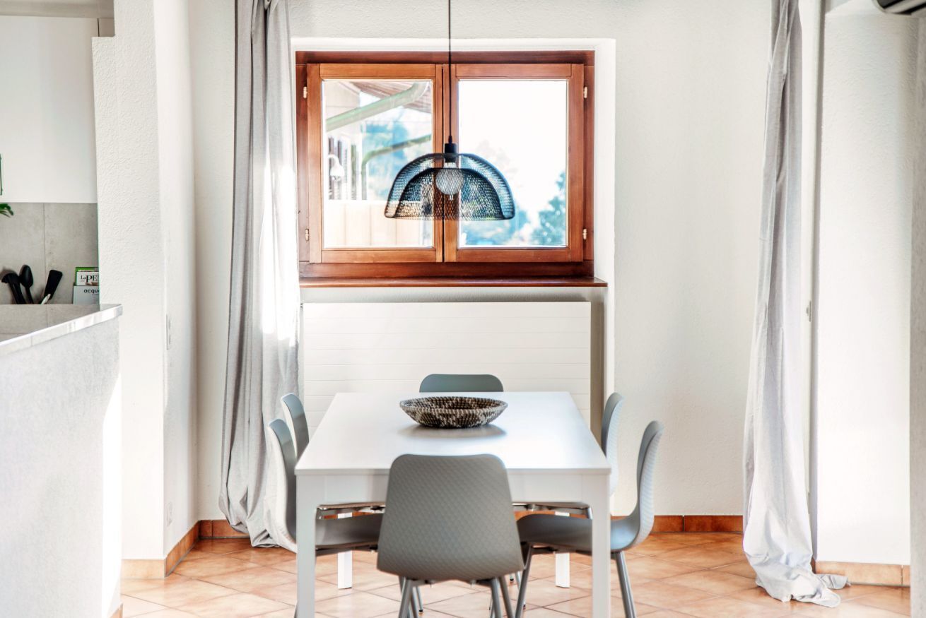 A dining area with a white table, grey chairs, a woven pendant light, and a window with curtains.