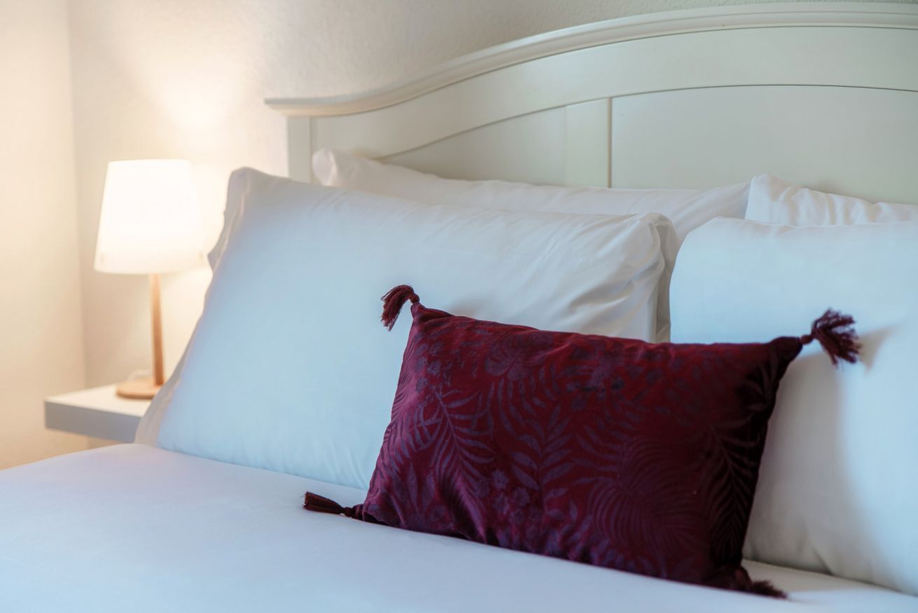 A neatly made bed with white and red pillows, a white headboard, and a bedside table with a lamp.