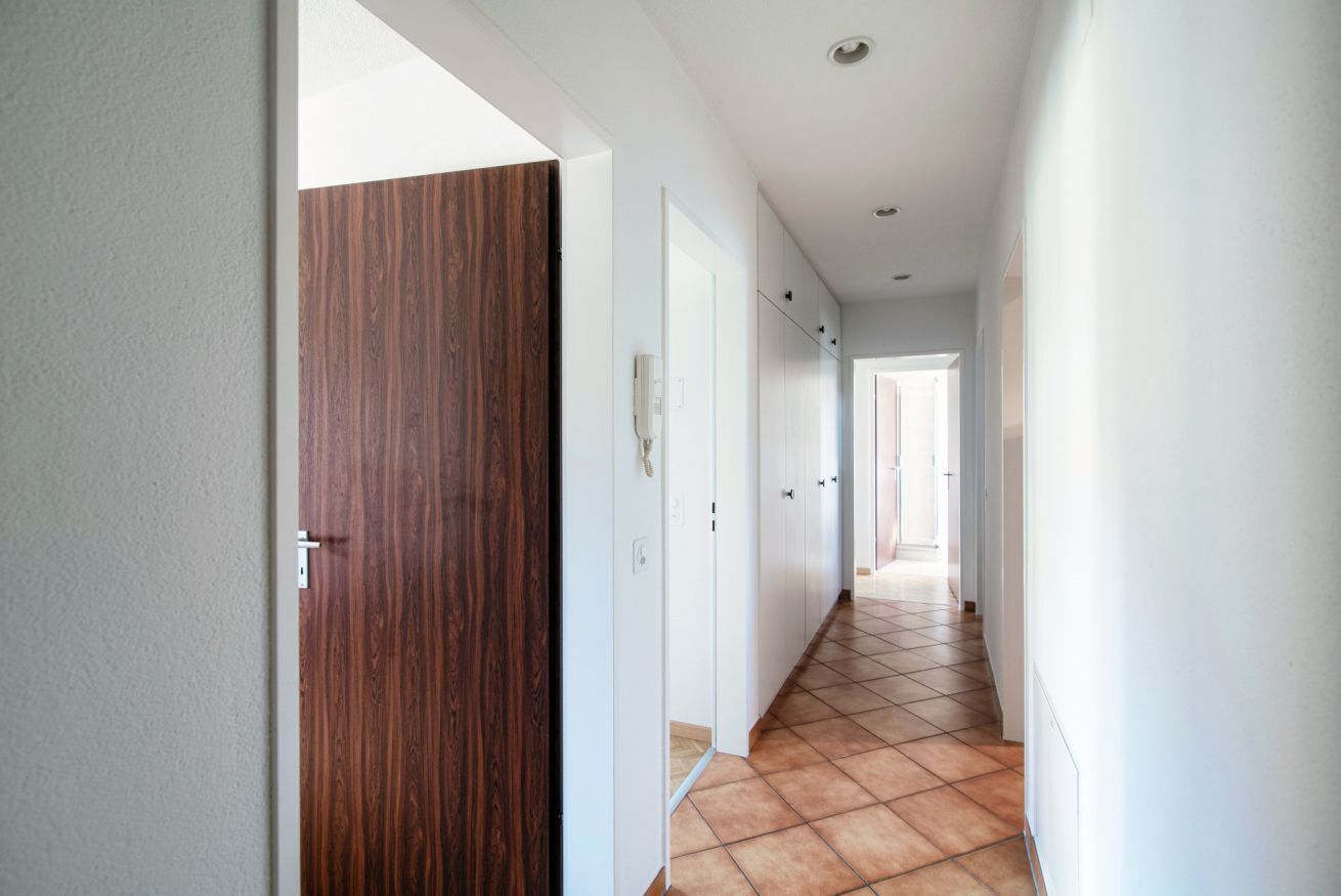 Un corridoio con una porta in legno, pareti bianche e pavimento piastrellato.