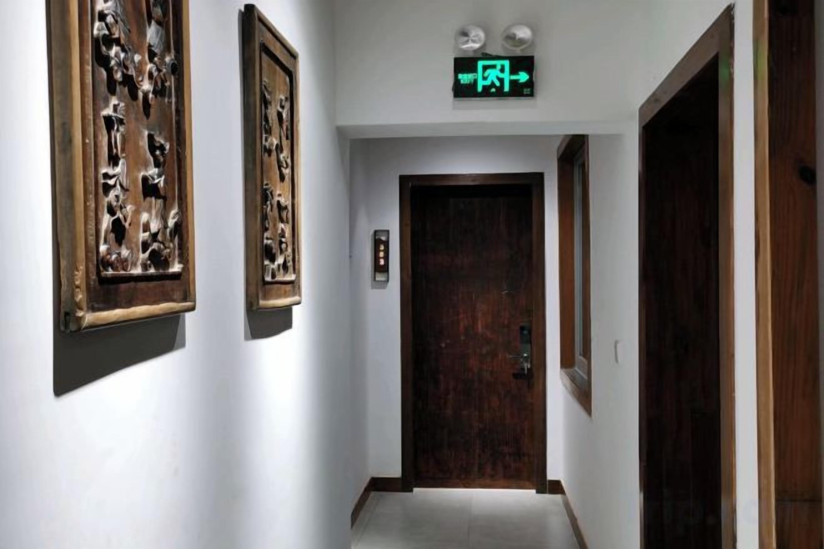 A hallway with a wooden door at the end, framed artwork on the walls, and a green exit sign above.