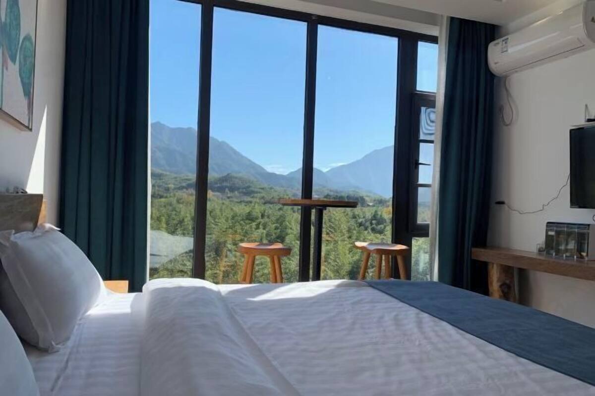 Ein Hotelzimmer mit einem großen Bett, einem Fernseher, einem Tisch, Stühlen und Blick auf Berge durch große Fenster.