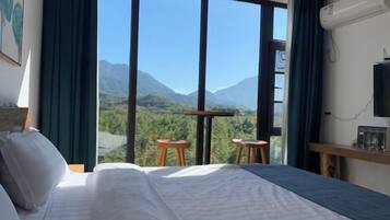 A hotel room with a large bed, a TV, a table, and chairs, and a view of mountains through large windows.