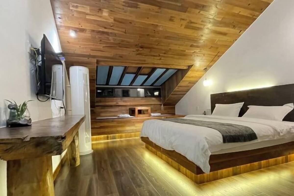 Ein Schlafzimmer mit schrägem Dach, Holzboden, einem Bett mit weißer Bettwäsche und einem kleinen Tisch mit einer Pflanze.