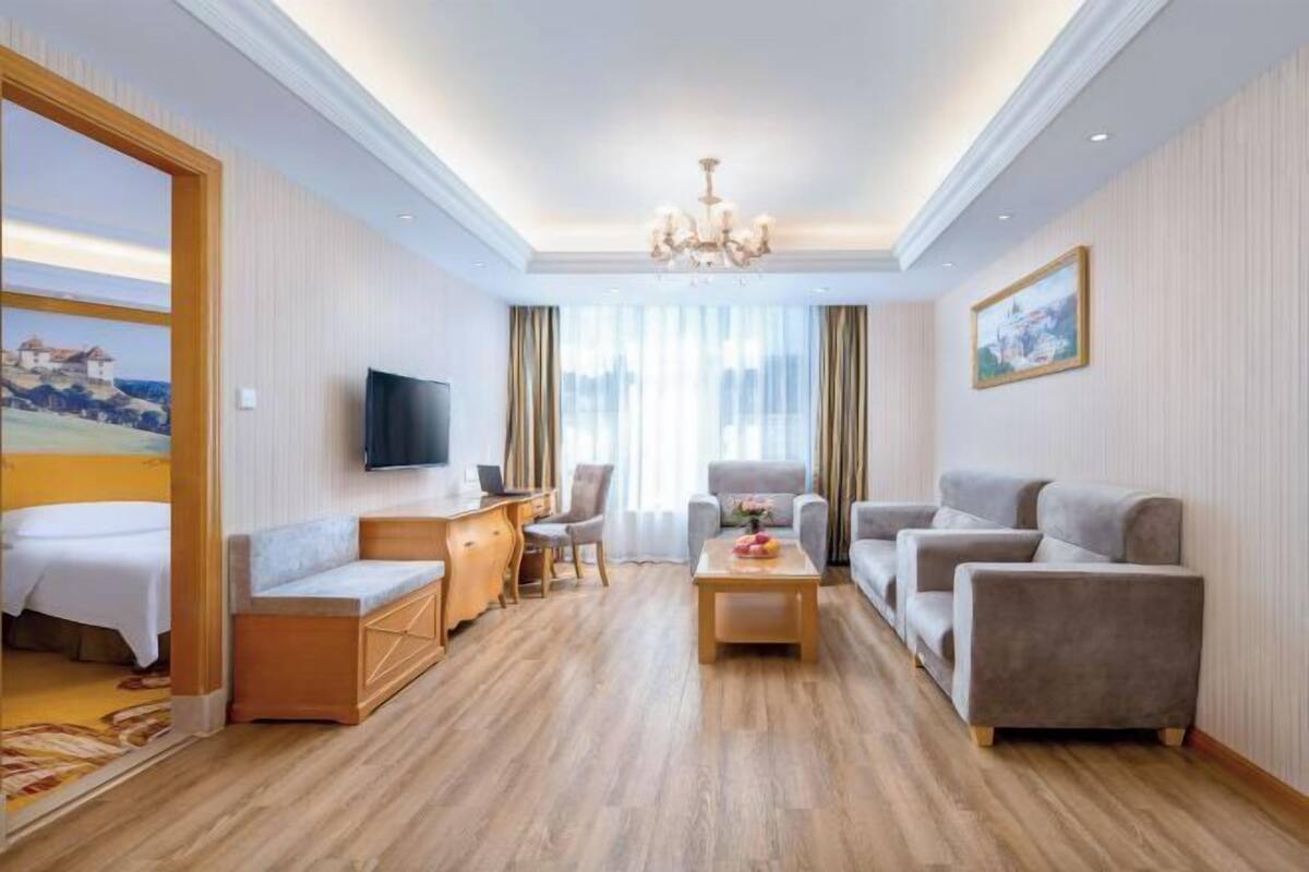 Habitación de hotel con piso de madera, televisor, escritorio con silla, un conjunto de sofás y un cuadro en la pared.