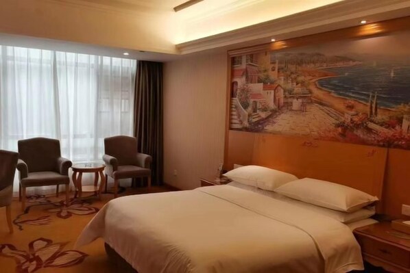 A hotel room with a large bed, two armchairs, a small table, and a painting of a coastal scene.