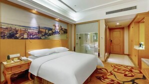 A hotel room with a large bed, a nightstand, a desk, a painting on the wall, and a door leading to a bathroom.