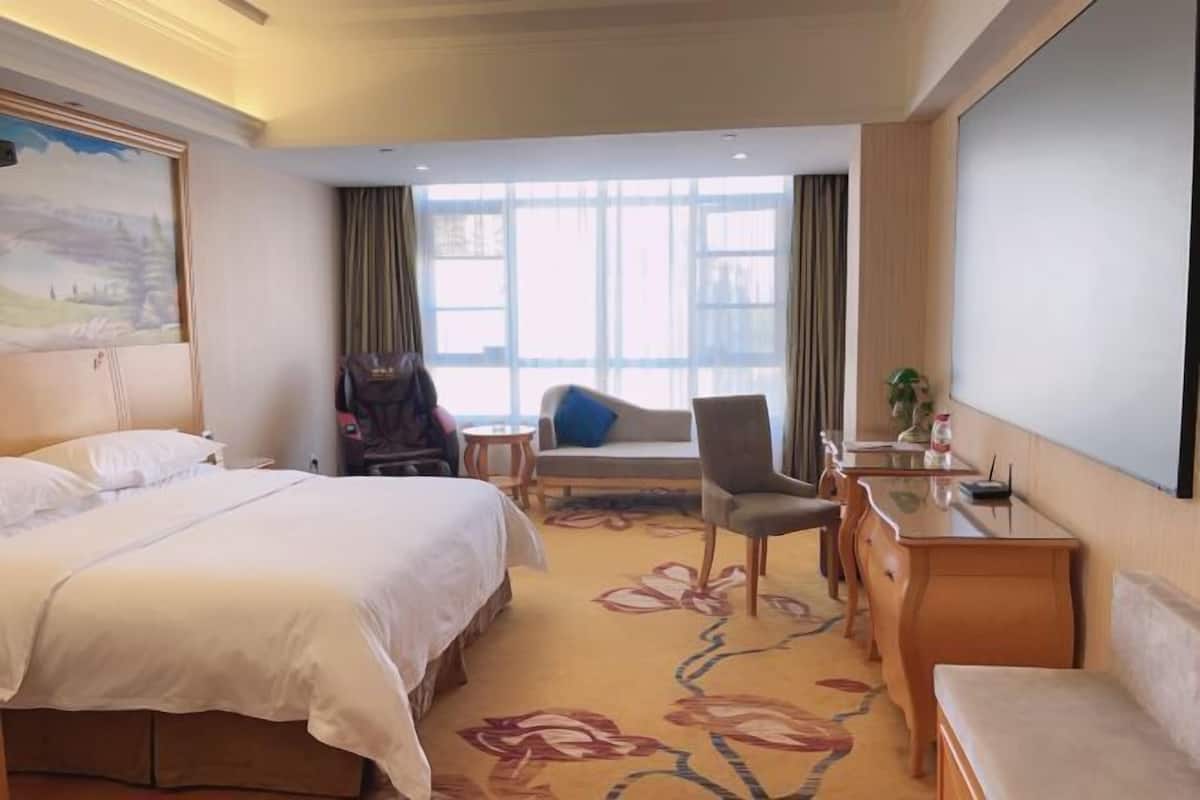 Habitación de hotel con una cama grande, un escritorio con silla, una mesita pequeña y un sofá. Hay una ventana con cortinas, un cuadro en la pared y vistas de la ciudad.