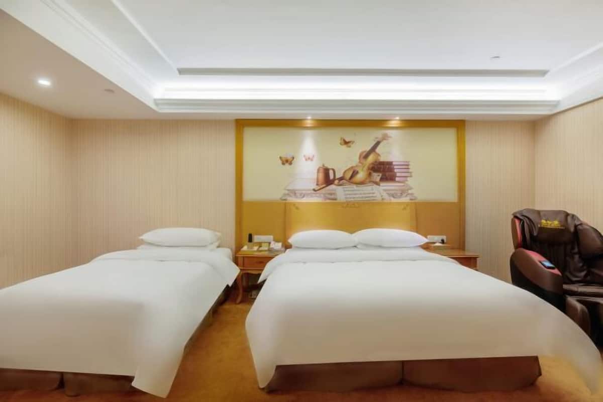 Habitación de hotel con dos camas individuales, cabecero con panel decorativo, sillón de masaje y televisor montado en la pared.