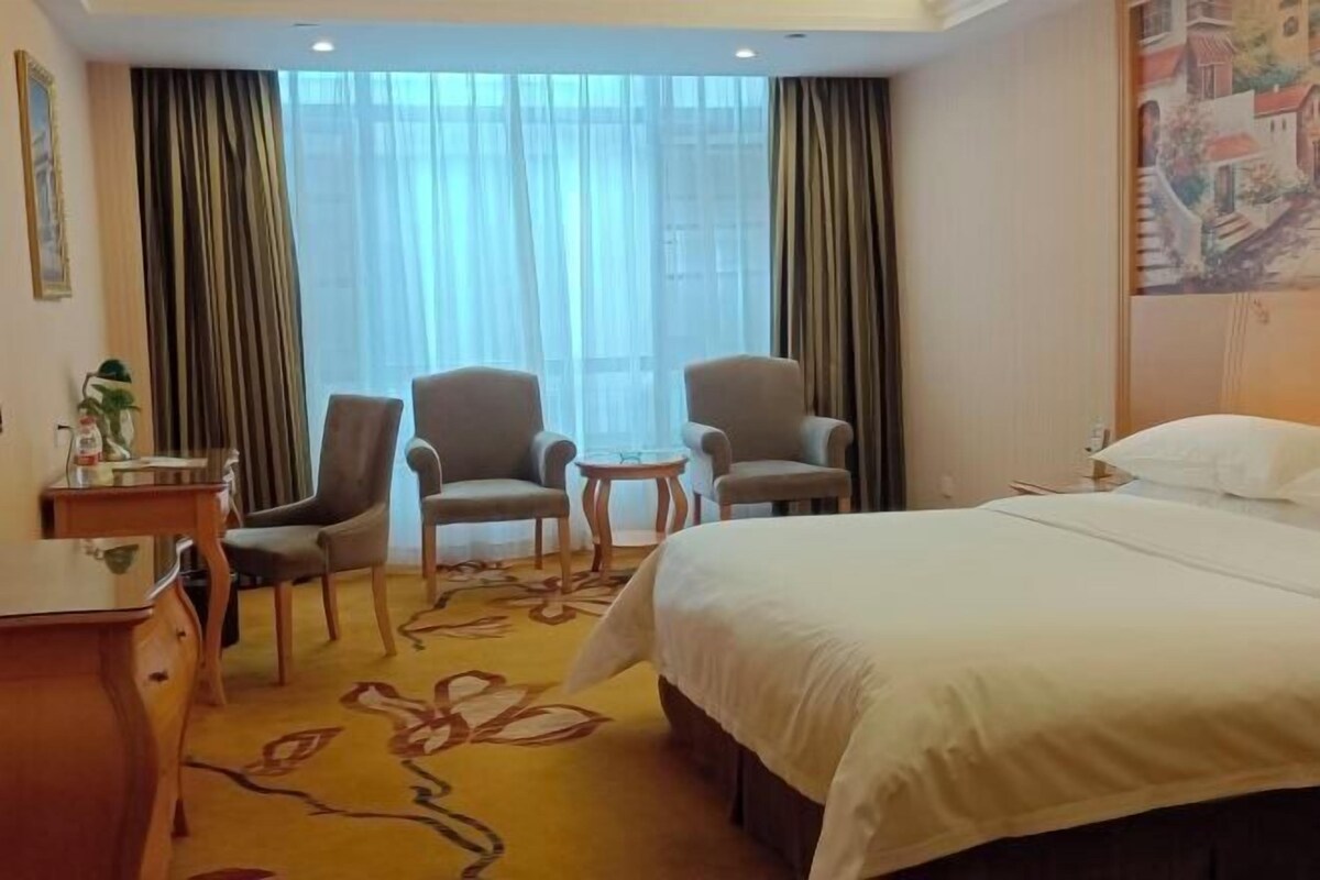 Hotellihuone, jossa on sänky, kaksi nojatuolia, pieni pöytä ja työpöytä.