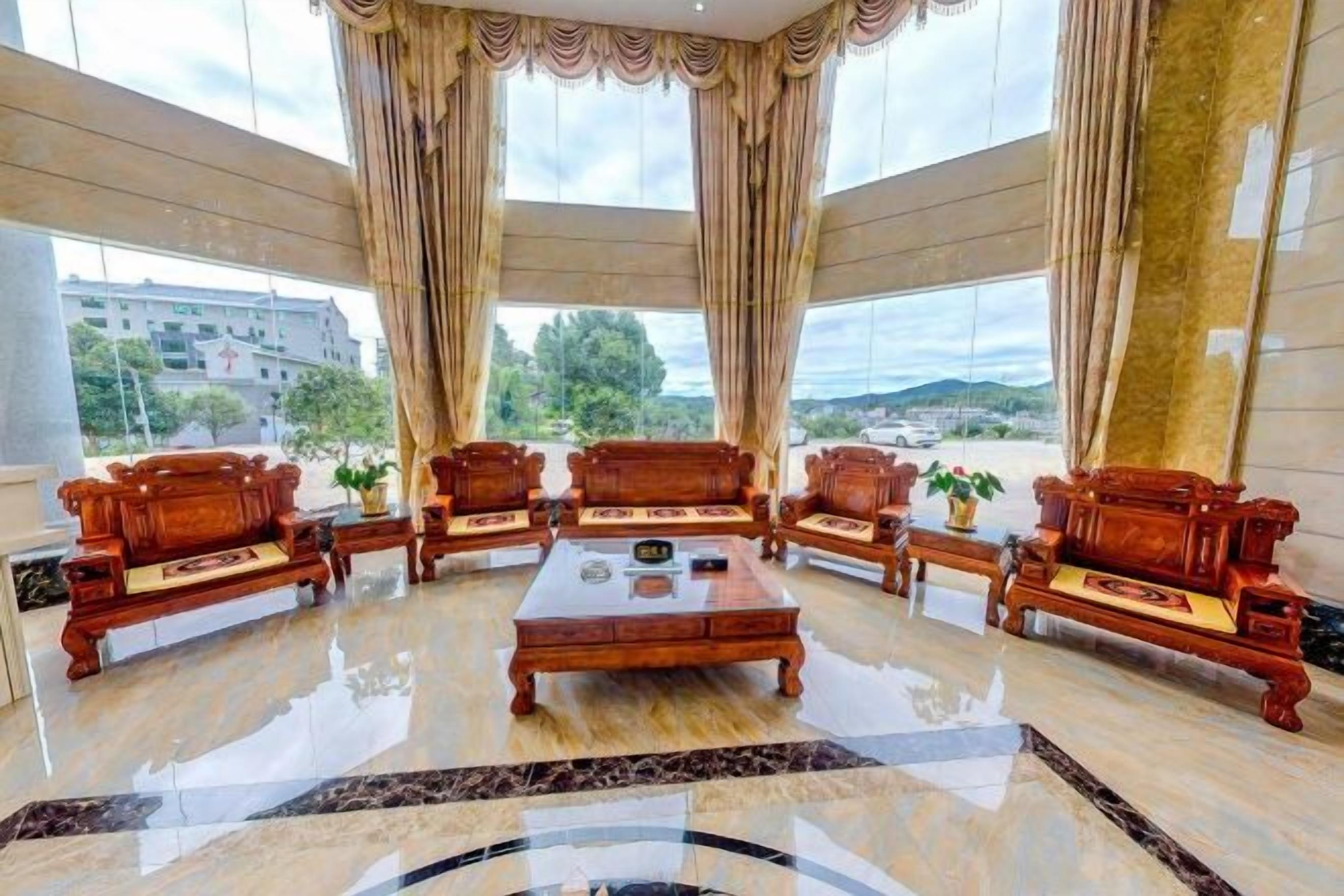A spacious lobby with a central coffee table surrounded by wooden sofas, large windows with curtains, and a view of the outside.