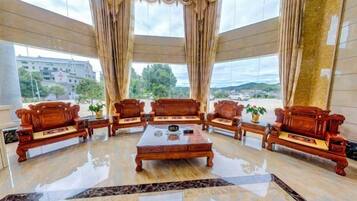 A spacious lobby with a central coffee table surrounded by wooden sofas, large windows with curtains, and a view of the outside.