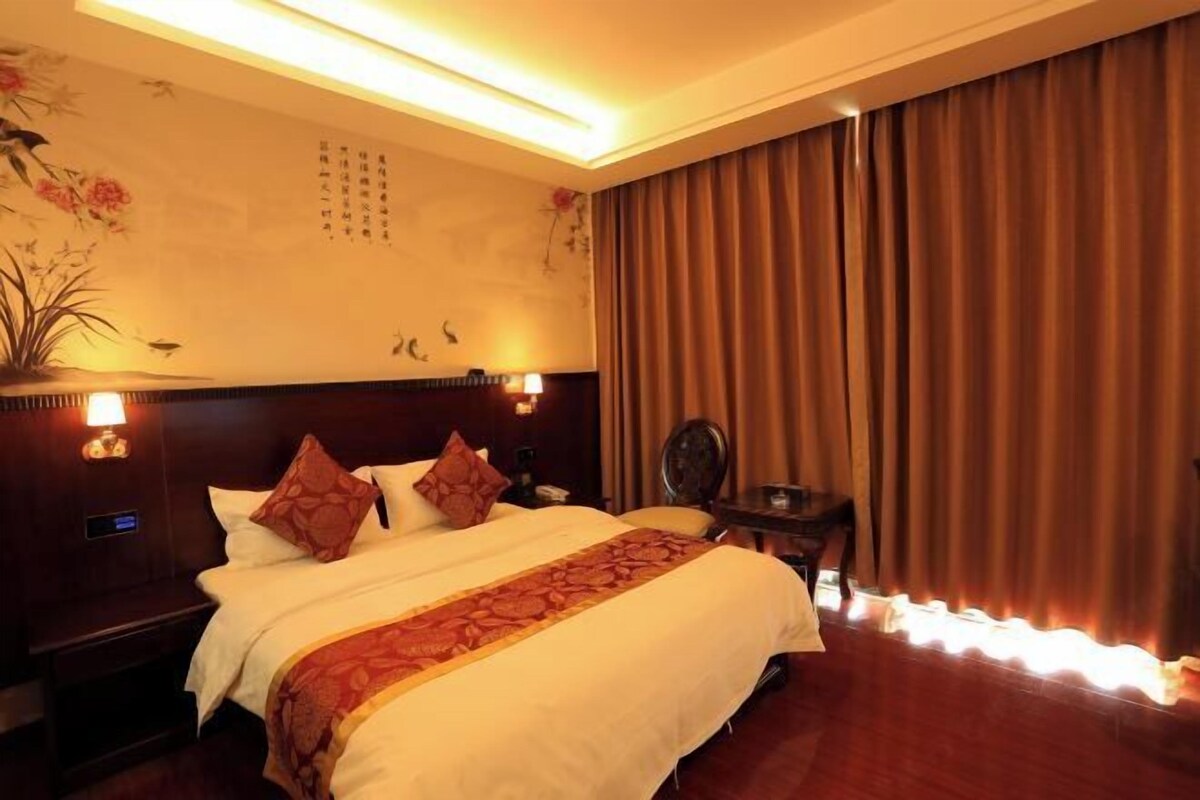 A hotel room with a large bed, a nightstand with a lamp, a wall with calligraphy, and a curtain.