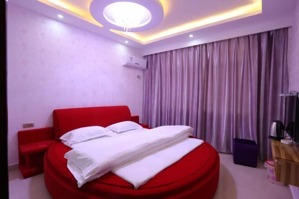 Una habitación de hotel moderna con una cama roja, un pequeño rincón de descanso, una lámpara de techo y una ventana con cortinas.