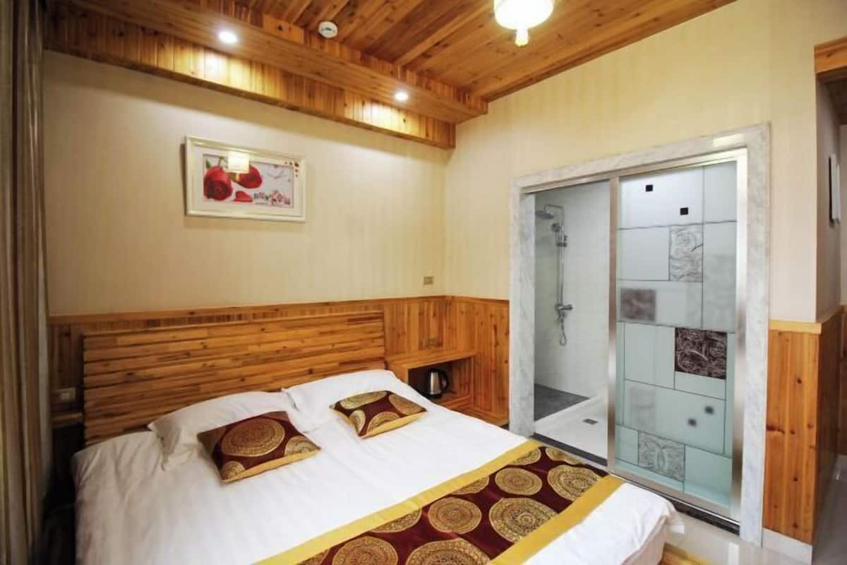 Un dormitorio con cabecera de madera, cama con ropa de cama estampada, una mesita de noche pequeña y una zona de ducha con paredes de azulejos.
