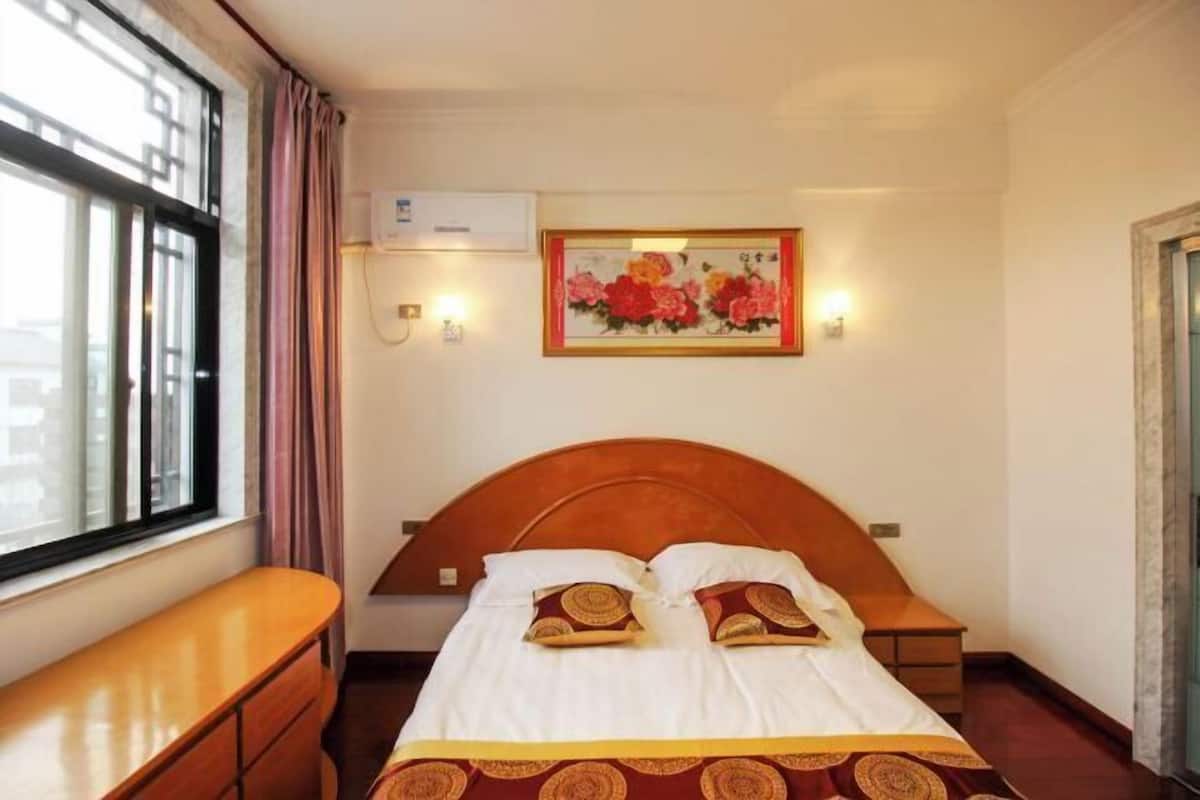 Habitación de hotel con una cama, una mesita de noche de madera, un escritorio, una ventana con cortinas, un equipo de aire acondicionado y un cuadro con flores en la pared.