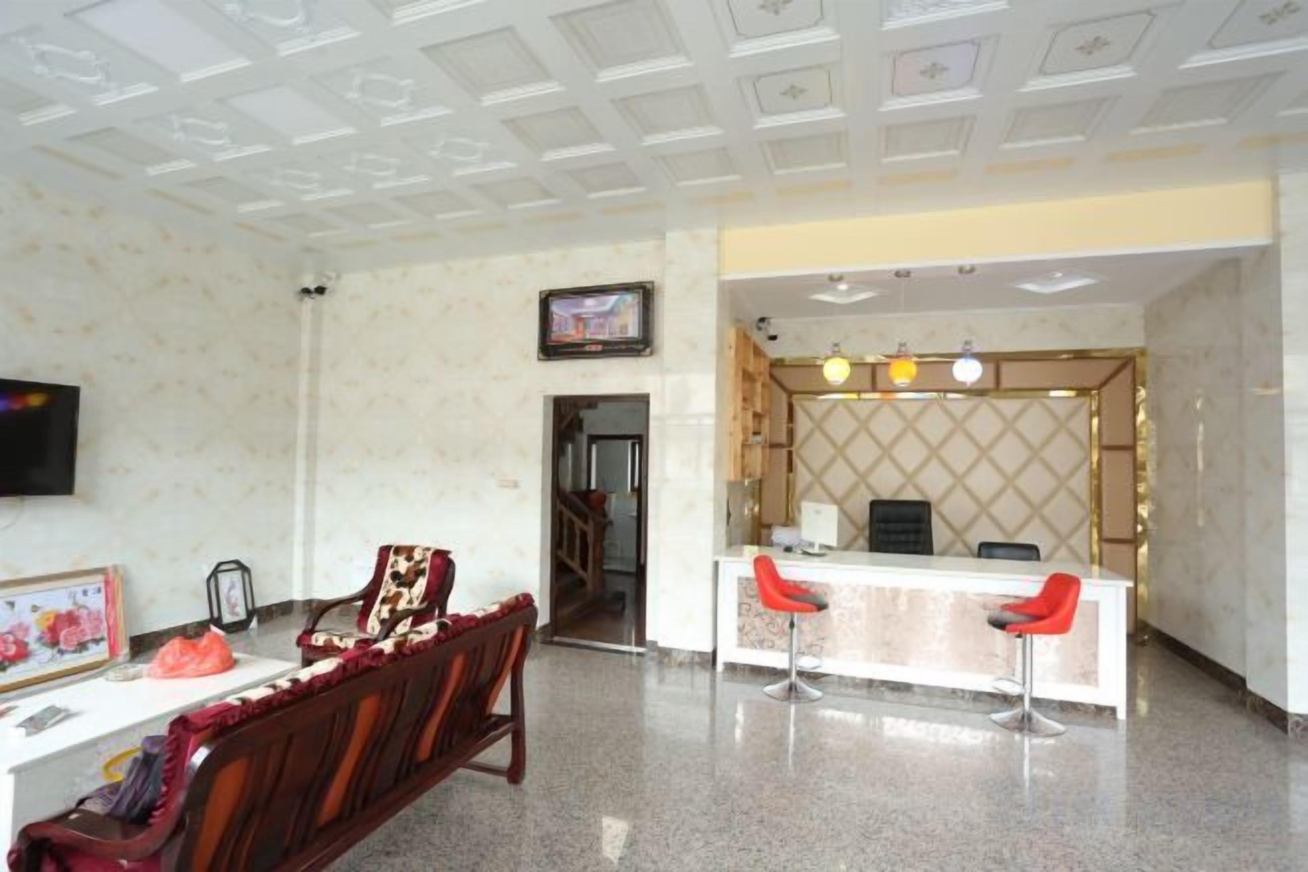 Een hotel lobby met een receptiebalie, twee rode barkrukken, een houten bank en een aan de muur bevestigde televisie.