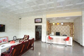 Eine Hotelhalle mit einer Rezeption, zwei roten Barhocker, einer Holzcouch und einem an der Wand montierten Fernseher.