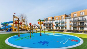 A playground with slides and climbing structures, surrounded by a pool with a blue and green design, and modern apartment buildings in the background.