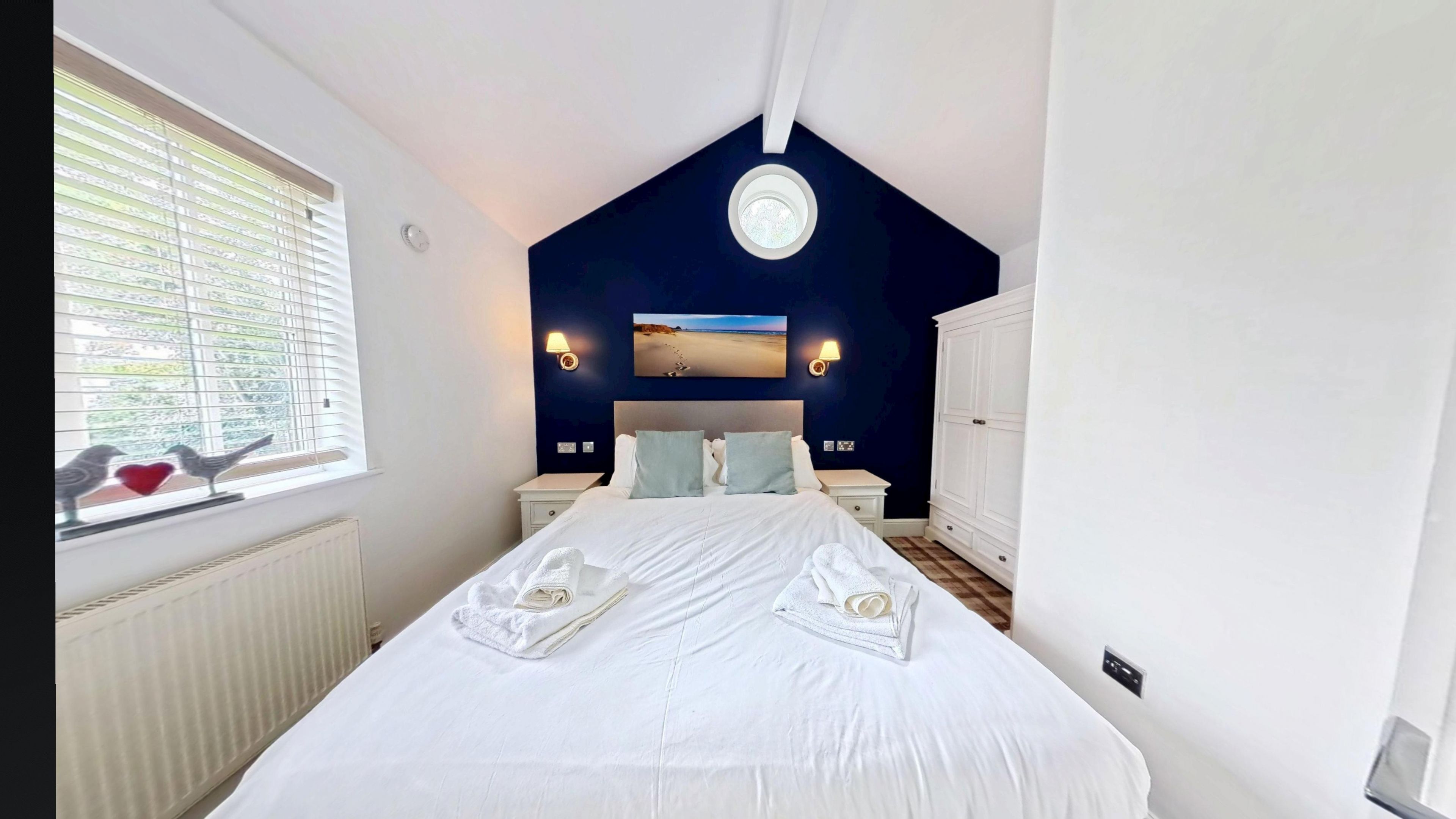A bedroom with a large bed, two bedside tables, a nightstand, a window with blinds, a wall-mounted picture, and a wardrobe.