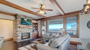 A spacious living room with a stone fireplace, a ceiling fan, and large windows offering a view of the outdoors. - Chalakee Lodge CJ-4- Mountain View, Gated Resort, Indoor/Outdoor Pools & Hot Tub (Boone)