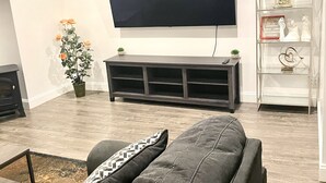 A living room with a flat-screen TV mounted on the wall, a grey sofa, a small table with a plant, and a decorative glass shelf. - Gardner 5 bedroom 3 bath home. Family friendly and long stay  (Gardner)