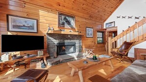 A cozy cabin interior with a stone fireplace, wooden walls, a mounted television, and a comfortable seating area. - Deer Ridge - 3BR, 3BA Cabin w/ Hot Tub, Firepit (Maggie Valley)