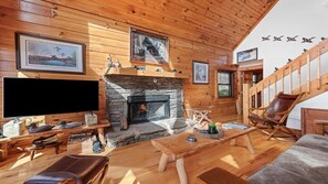 A cozy cabin interior with a stone fireplace, wooden walls, a mounted television, and a comfortable seating area.