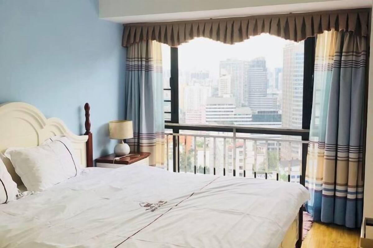 A hotel room with a large bed, a nightstand, a lamp, and a view of the city through a balcony.