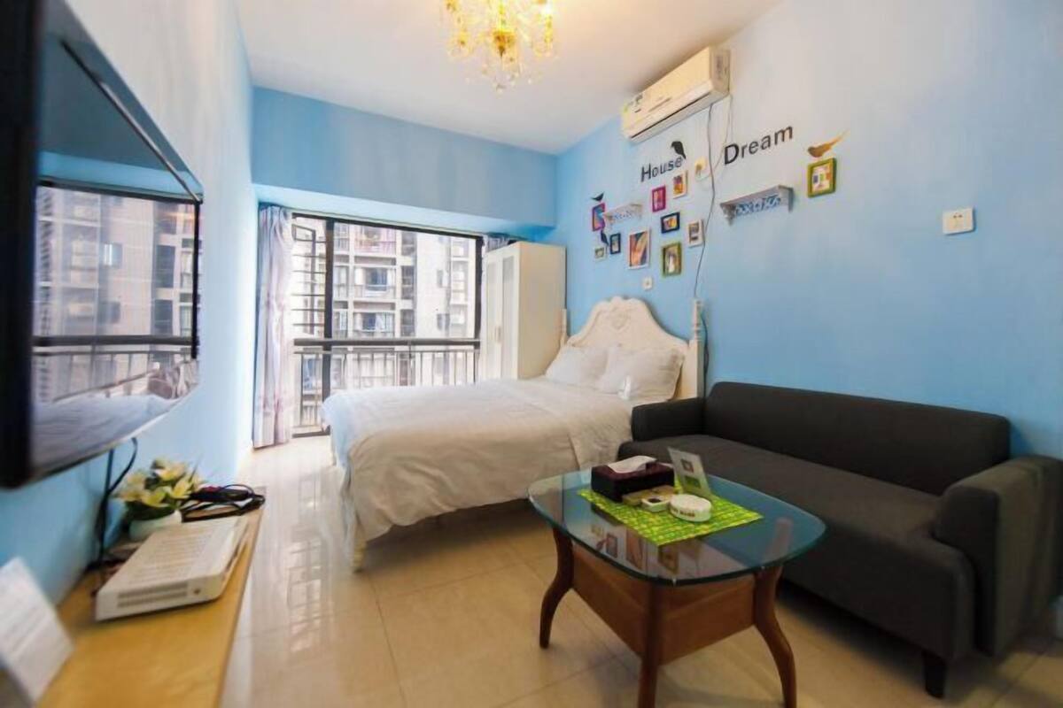 A bedroom with a bed, a glass coffee table, a sofa, a TV, and a window with a view of buildings.