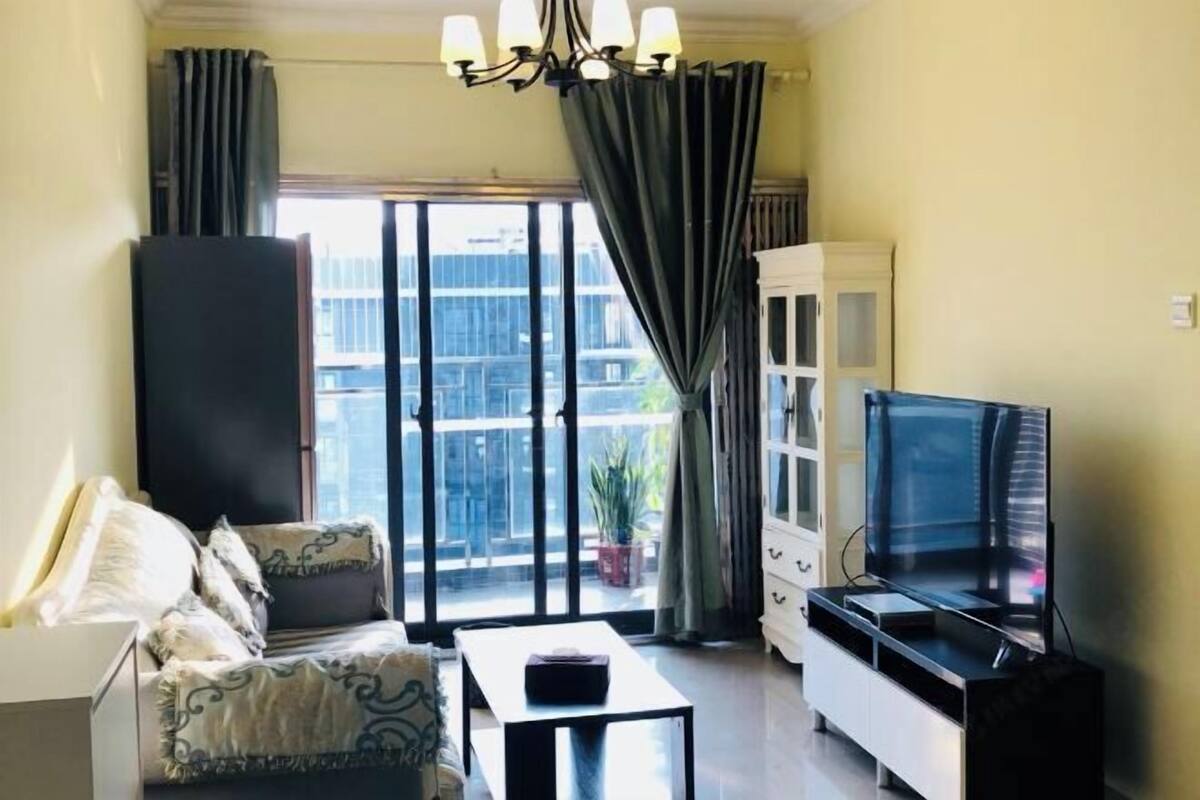 A living room with a sofa, a coffee table, a TV on a stand, a cabinet, and a balcony with curtains.