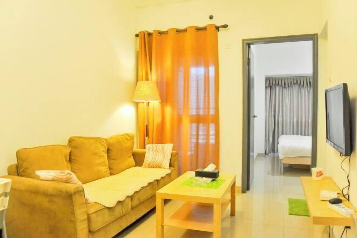 A room with a sofa, a table, a lamp, a television, and a bed in the background.