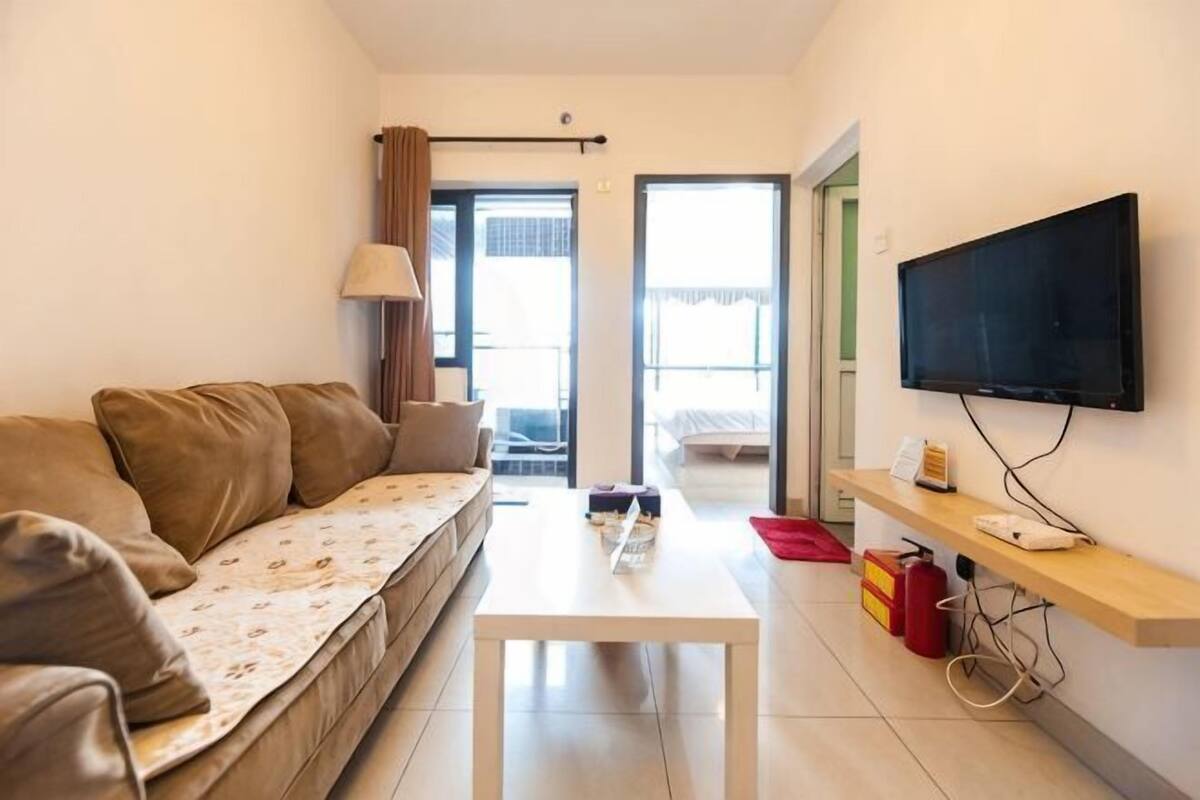 A living room with a sofa, a coffee table, a flat-screen TV, and a small shelf with a phone and a red object.