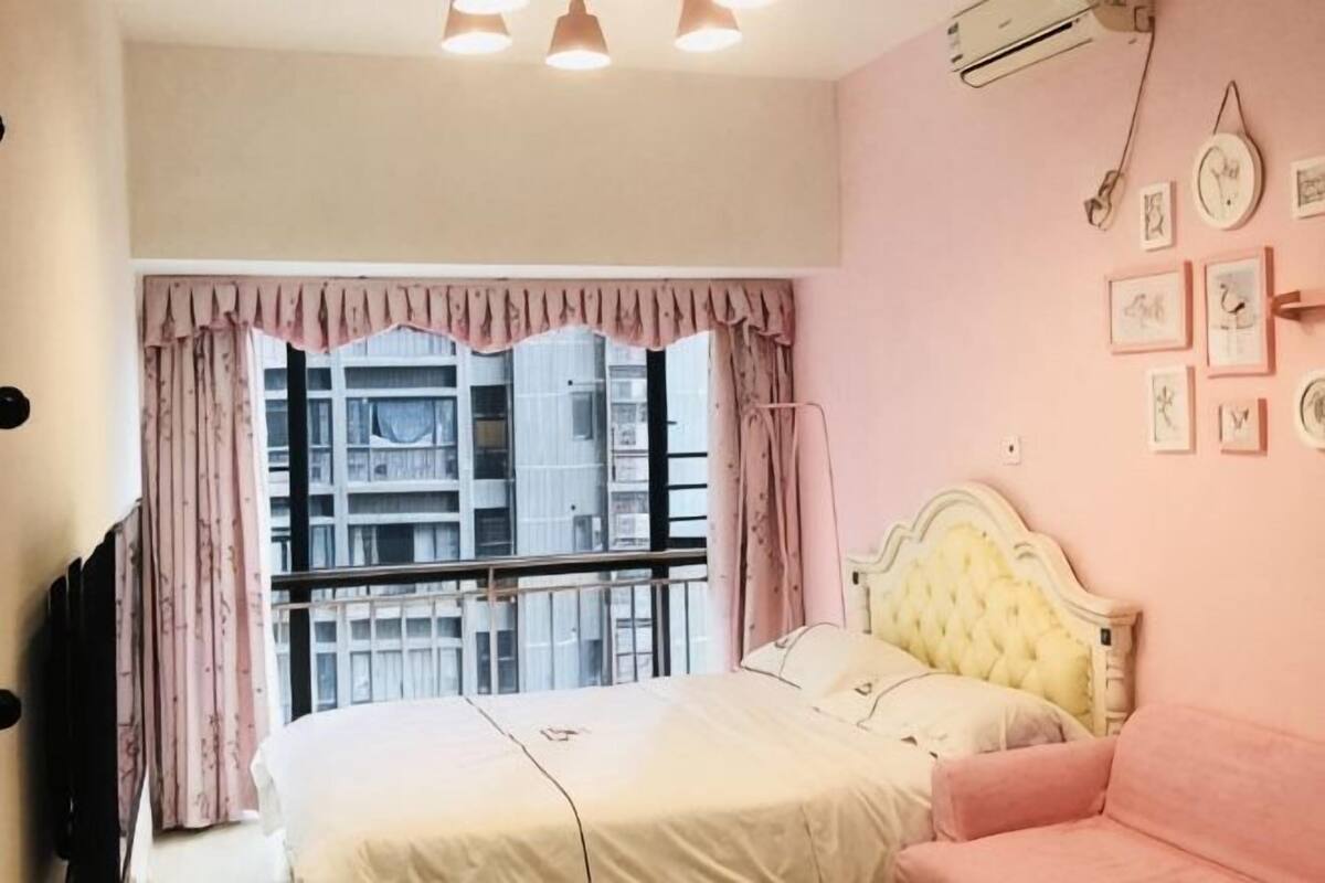 A bedroom with a bed, a pink sofa, a window with curtains, and a wall with framed pictures.