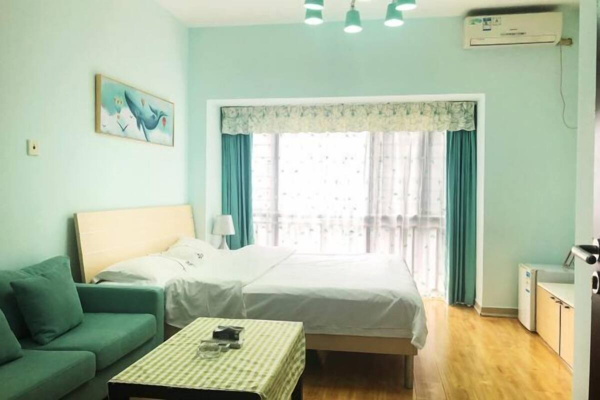 A room with a bed, a sofa, a table, a window with curtains, and an air conditioning unit.