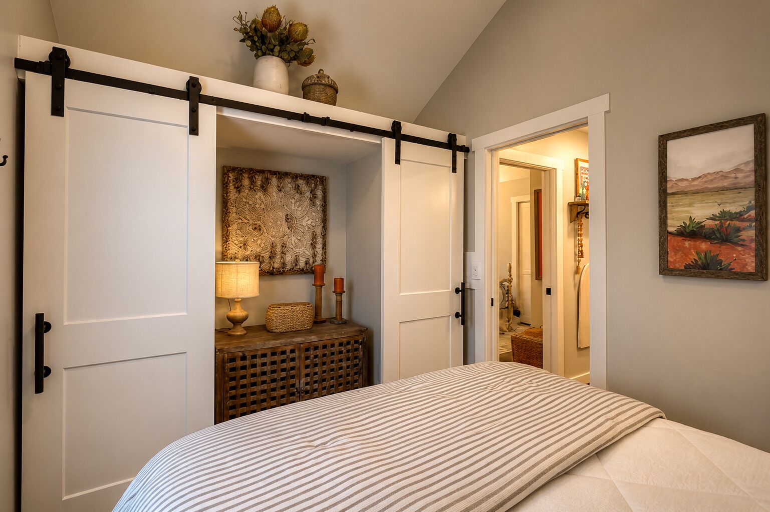 Un dormitorio con cama, mesita de noche, lámpara, un adorno en la pared y una puerta corredera que da a otra habitación.