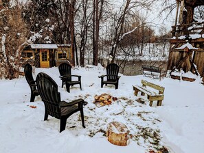 Lumipeitteinen ulkoalue, jossa on Adirondack-tyylisiä tuoleja ja puinen penkki. Alue on puitten ja puisen rakennelman ympäröimä.