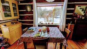 Un acogedor interior de cabaña con una mesa de comedor de madera, sillas, un armario con vajilla y una ventana con vistas a árboles.