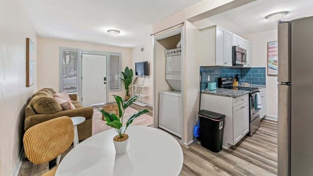 Un espace de vie compact comprenant un coin cuisine, un coin repas avec une table et des chaises blanches, un canapé et une plante en pot.
