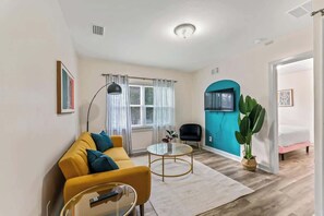 A modern living room with a yellow sofa, a glass coffee table, and a black chair. There is a large window with curtains, a teal accent wall, and a potted plant. - The Hendricks #4: Near San Marco (Jacksonville)
