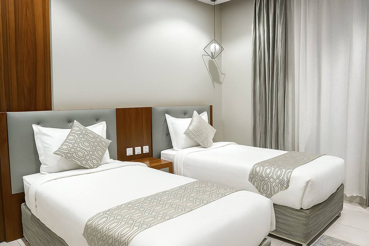 Habitación de hotel con dos camas, cabecera de madera, mesita de noche, lámpara de pared y ventana con cortinas.