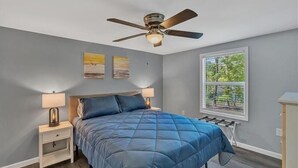 Une chambre à coucher avec un lit, deux lampes de chevet, un ventilateur de plafond et une fenêtre donnant sur des arbres.