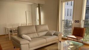 Ein modernes Wohnzimmer mit einem beigen Sofa, einem Glastisch und einem Essbereich mit weißen Stühlen.