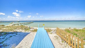 A wooden boardwalk leading to a beach with clear water and a distant pier.