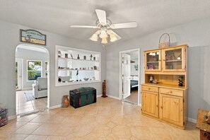 Una habitación amplia con un ventilador de techo, un mueble de madera con puertas de cristal y un estante de pared con varios objetos decorativos.