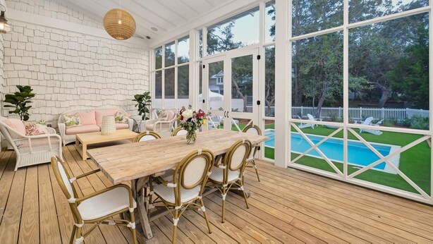 Un patio cubierto con mesa de madera y sillas de mimbre, con vistas a una piscina y una valla de piquetes blancos.