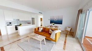 A modern living room with a sofa, coffee table, and dining area. There is a kitchen with white cabinets and a refrigerator. A painting is on the wall, and a window with curtains is visible. - Paraíso Benahavís - Private Garden Ground Floor (Benahavís)
