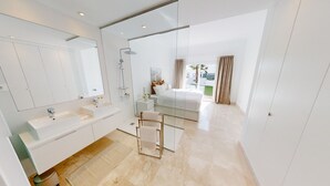 A modern hotel bathroom with a large mirror, two sinks, a shower, and a glass-enclosed bedroom with a bed and a view of the outside. - Paraíso Benahavís - Private Garden Ground Floor (Benahavís)