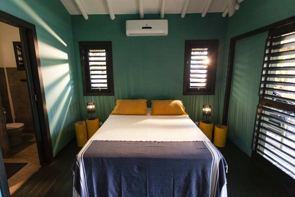 A bedroom with a bed, two bedside lamps, an air conditioning unit, and two windows with blinds.