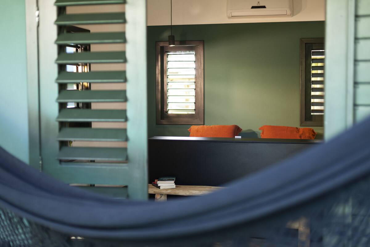 A room with a hammock, a bed with orange pillows, a window with blinds, and a small table with books.