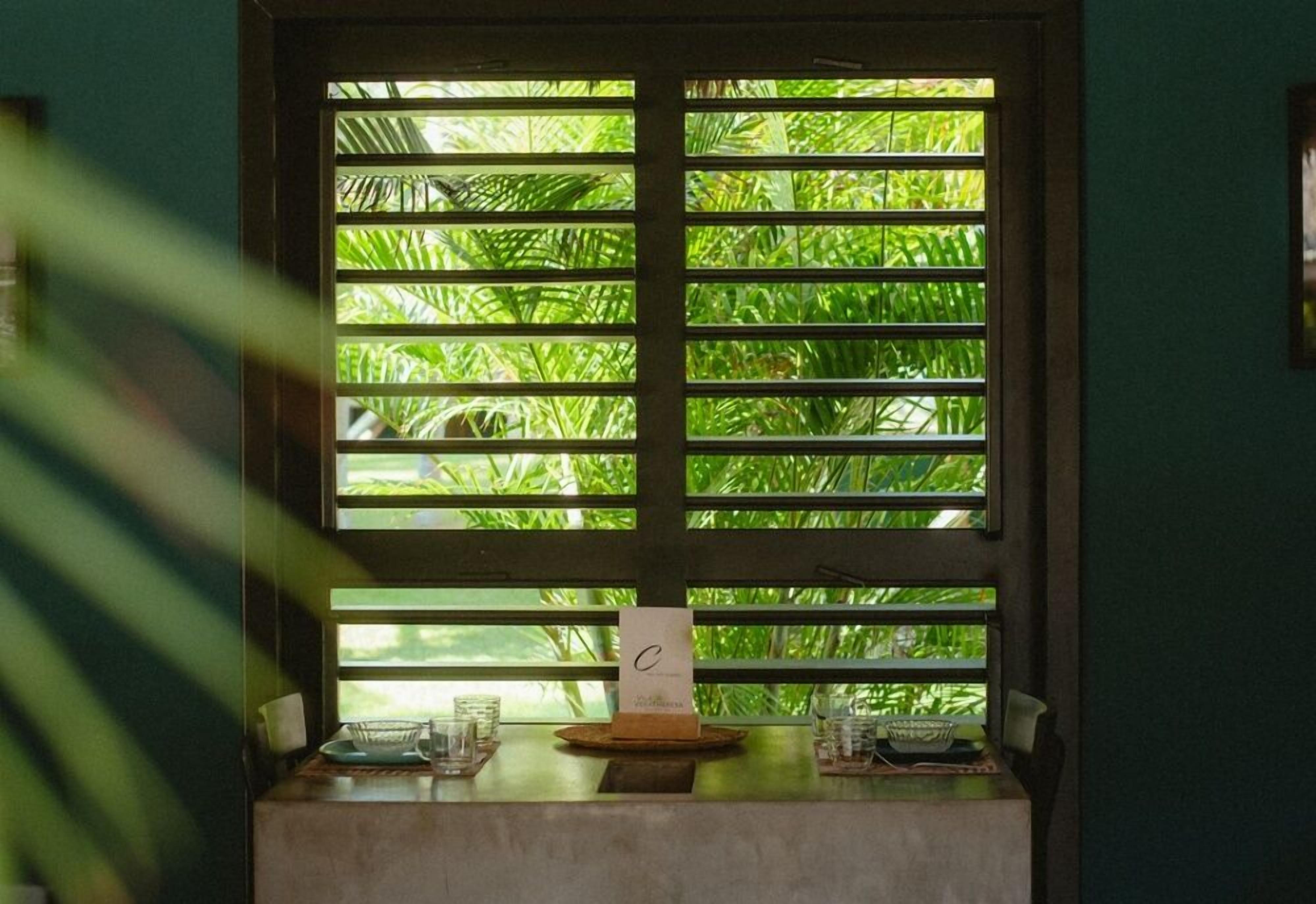 A window with horizontal blinds, a small table with a menu stand, and a view of lush green foliage outside.